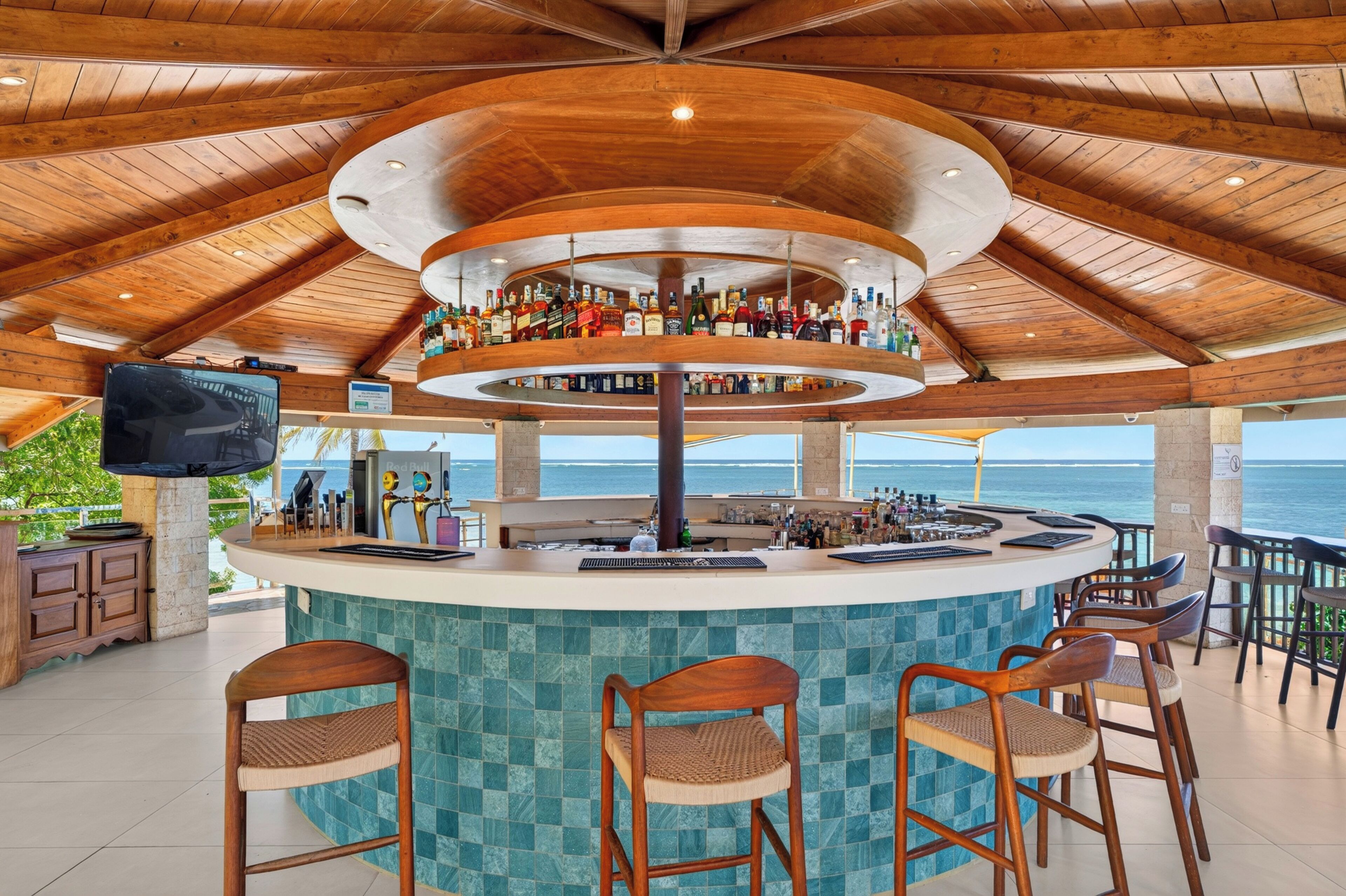 A bar with a circular design, multiple liquor bottles, a television, and a view of the ocean.