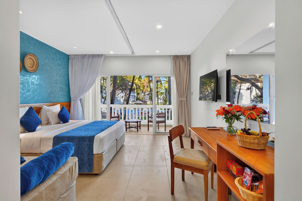 Une chambre d’hôtel avec un lit, un bureau, une chaise, une télévision et un balcon donnant sur des palmiers.