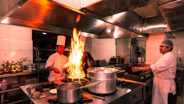 A professional kitchen with chefs working, a large stove with a pot on fire, and various cooking utensils and equipment. - Kathmandu Eco Hotel (Kathmandu)