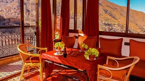 Uma varanda com cadeiras de vime, uma mesa com toalha de mesa vermelha e vista para uma aldeia montanhosa.