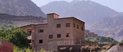 A two-story building with multiple windows, surrounded by greenery and mountains in the background.