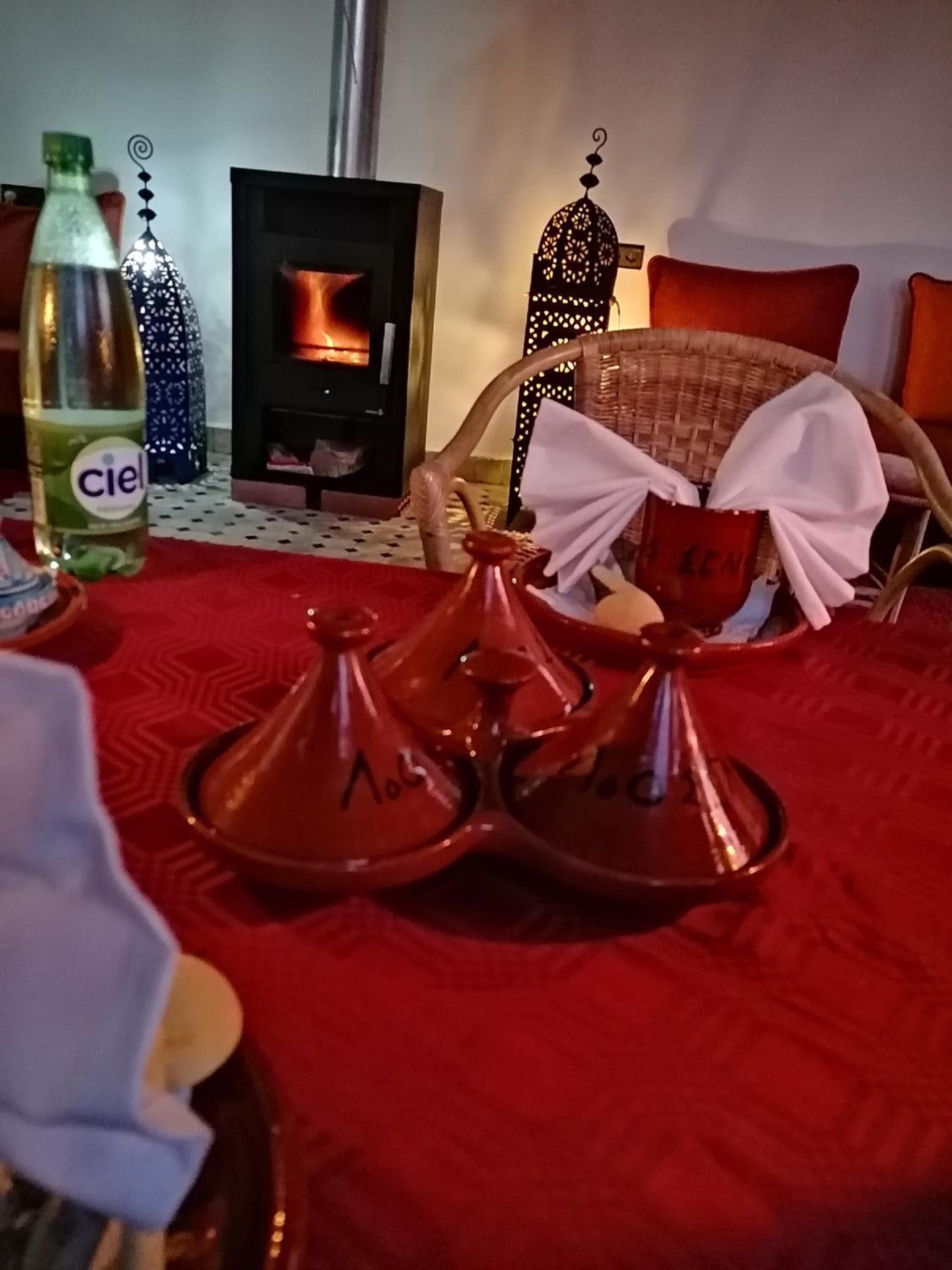 Una habitación acogedora con alfombra roja, una botella de agua Ciel, un juego de té marroquí tradicional, una cesta tejida y una chimenea encendida.