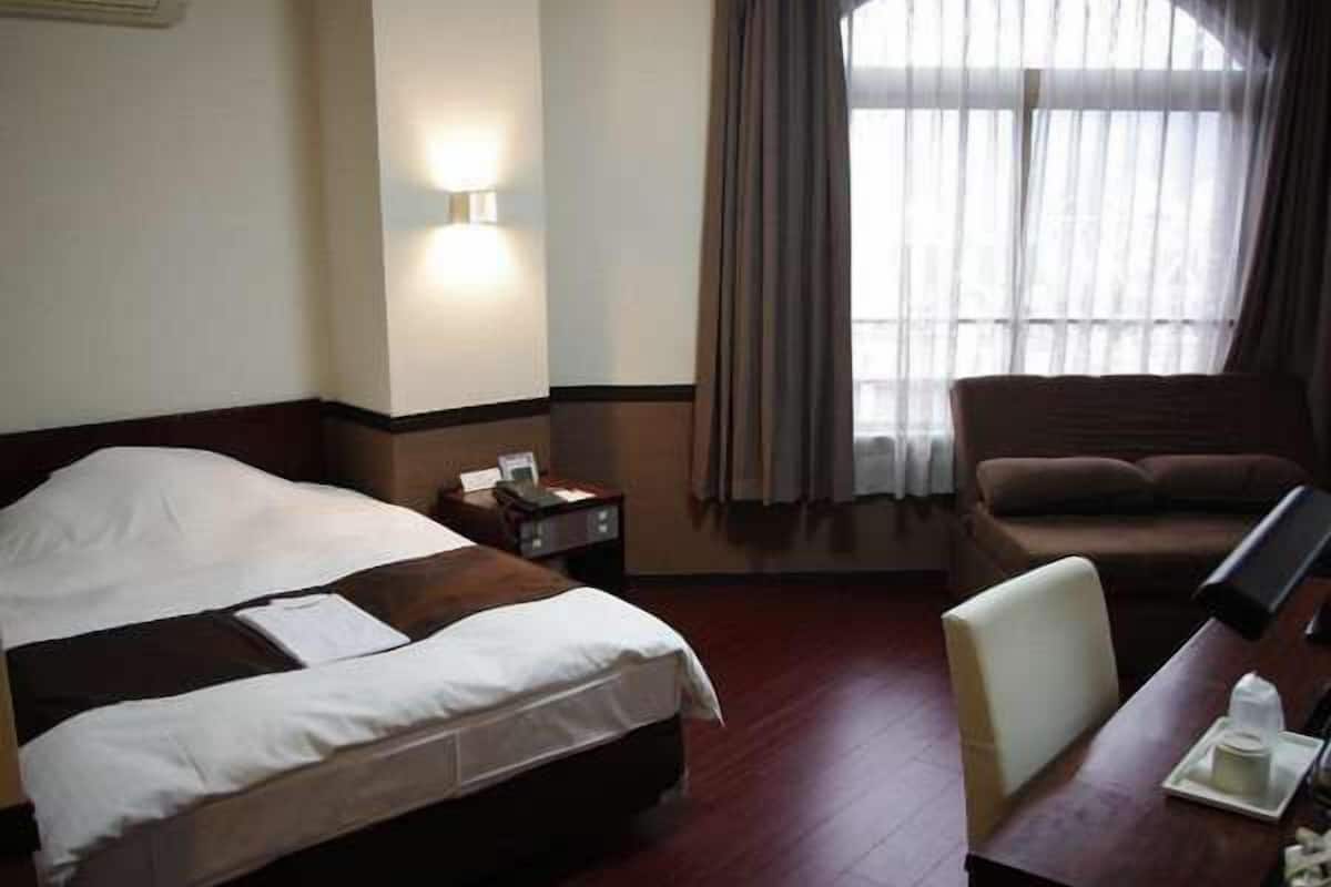 A hotel room with a bed, a desk, a chair, a nightstand, and a window with curtains.