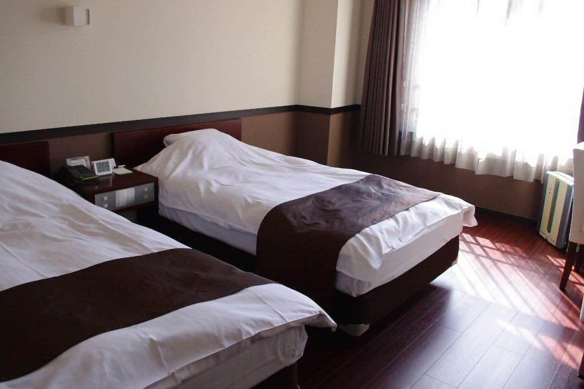 A hotel room with two beds, a nightstand, a bedside table, and a window with curtains.
