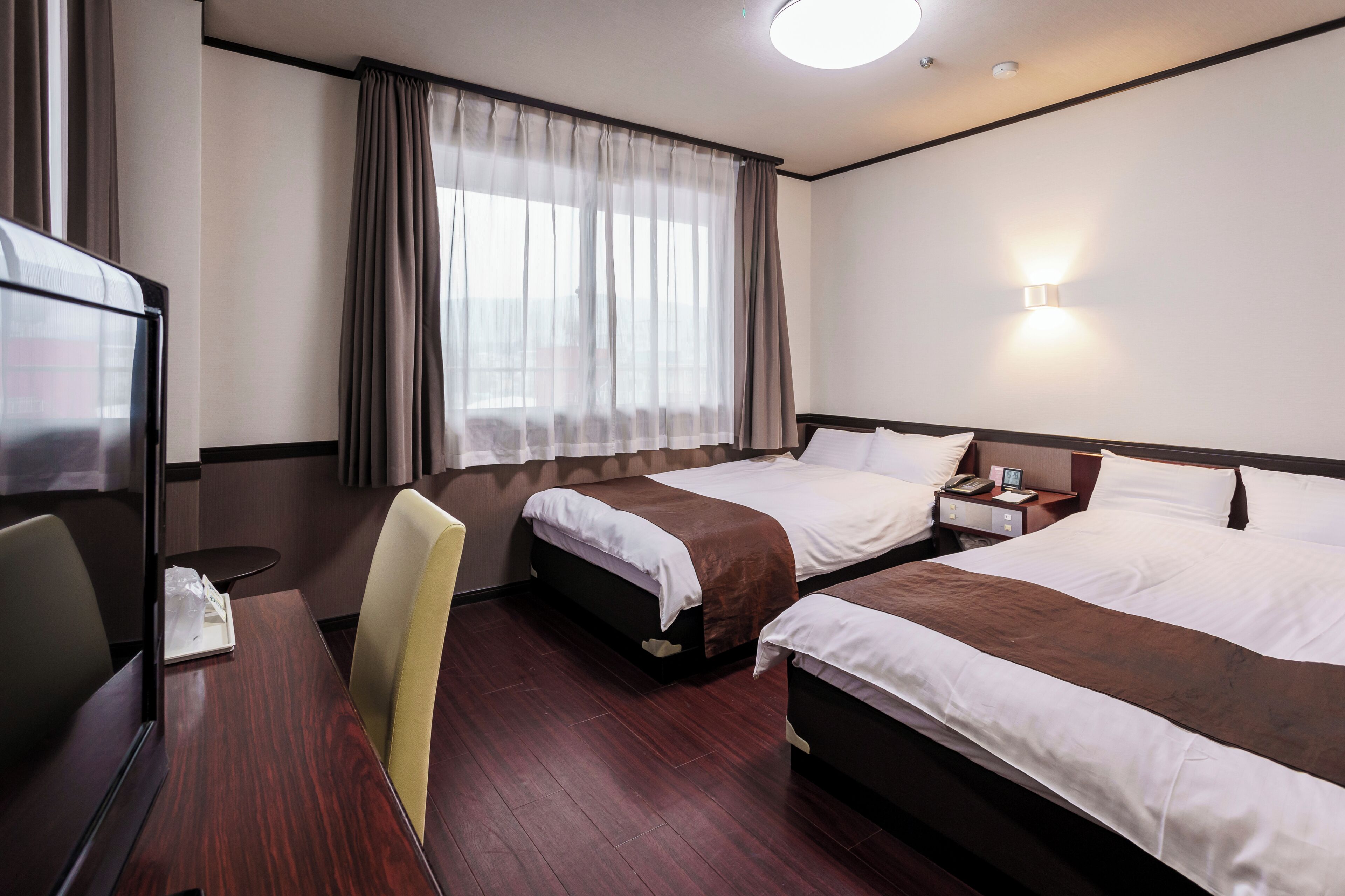 Una habitación de hotel con dos camas, un escritorio con silla, televisión y ventana con cortinas.