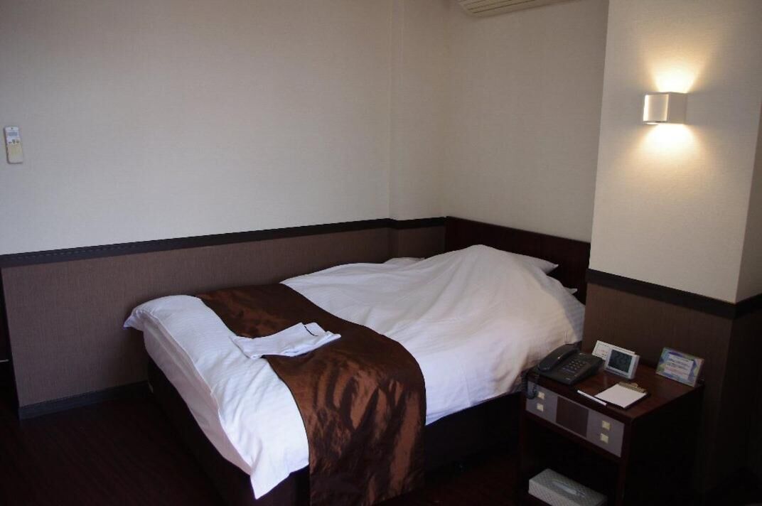 Habitación de hotel con una cama individual, una mesita de noche con un teléfono y un reloj pequeño, y una lámpara fijada a la pared.