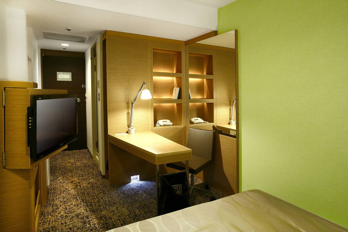 A hotel room with a bed, a desk with a lamp, a television, a telephone, and a trash bin.
