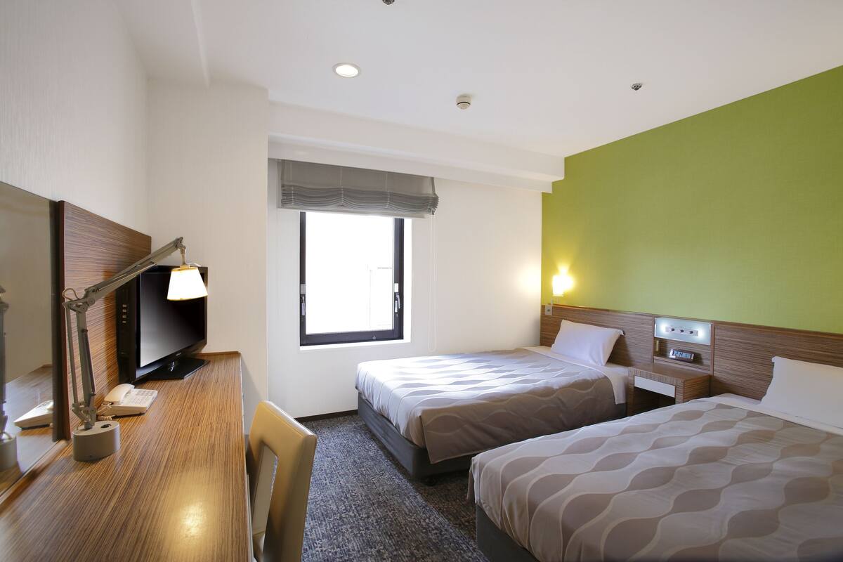 A hotel room with two beds, a desk with a computer and lamp, a television, and a window with a view.