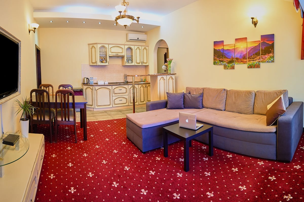 A living room with a sofa, a coffee table, a TV, and a kitchen in the background.