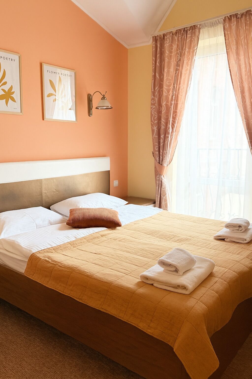 A hotel room with a bed, pillows, a headboard, a wall-mounted lamp, and curtains.