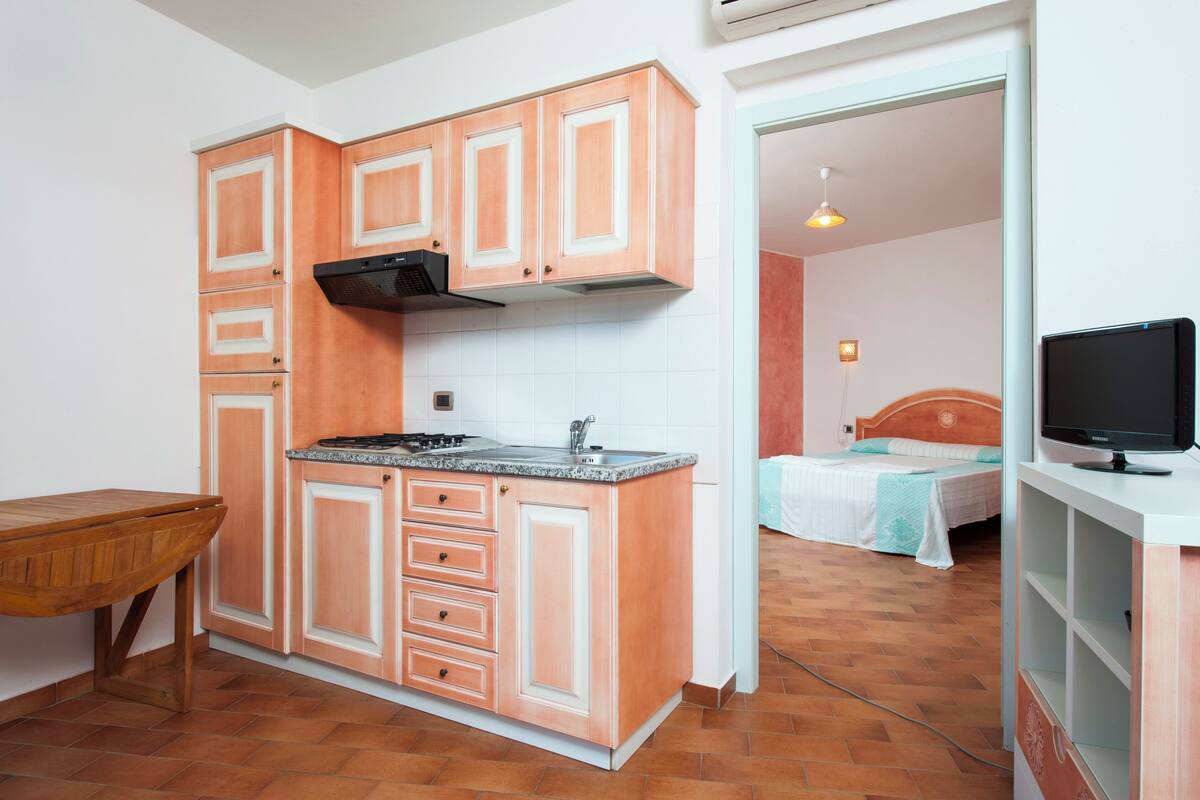 A compact kitchen with wooden cabinets, a countertop, and a stove. There is a bed with a headboard in the adjacent room.