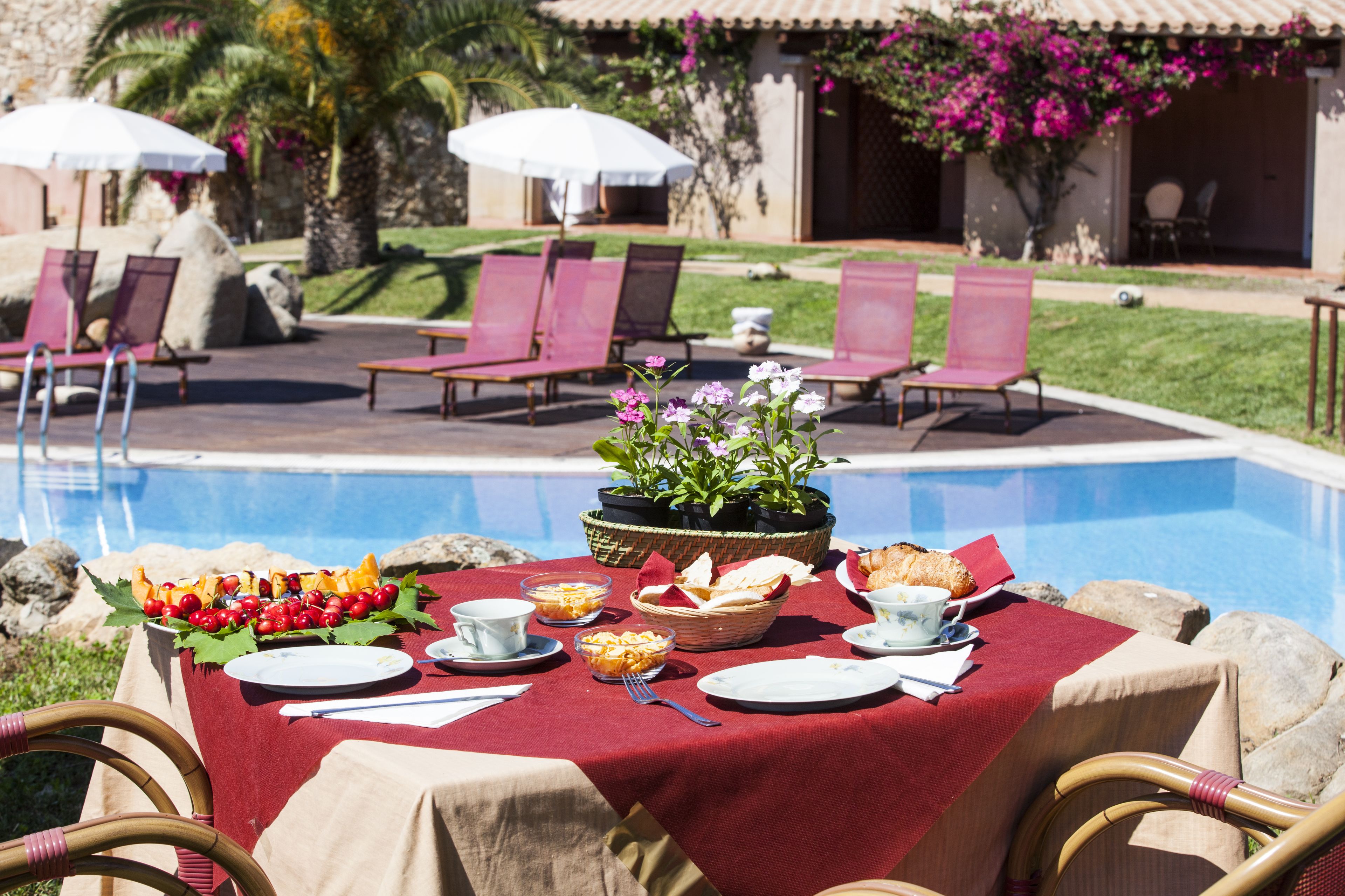 A poolside dining setup with a table covered in a red tablecloth, surrounded by lounge chairs and a pool.