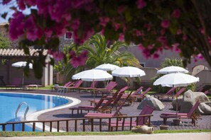 A pool area with lounge chairs and umbrellas, surrounded by palm trees and a stone building. - S'Incantu Resort (Villasimius)