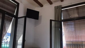 Un dormitorio con cama, una mesita de noche de madera, televisor montado en la pared y ventanas amplias con persianas.