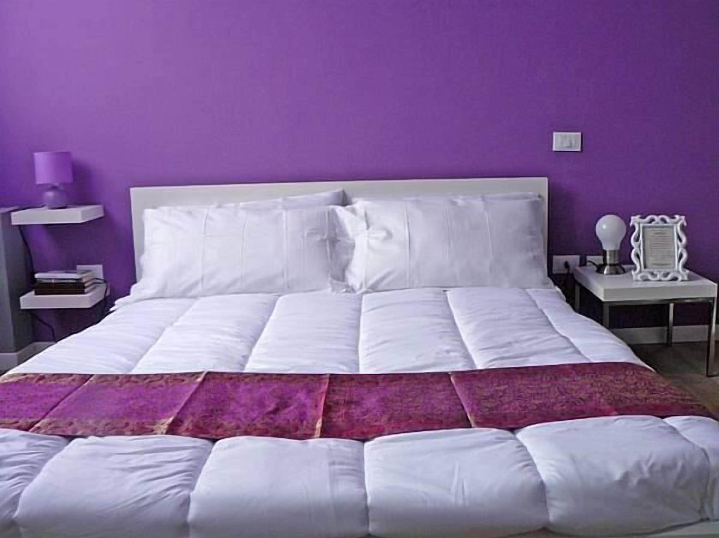 A bedroom with a large bed, white bedding, a purple wall, and a nightstand with a lamp.