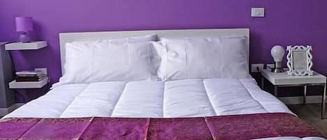 A bedroom with a large bed, white bedding, a purple wall, and a nightstand with a lamp.