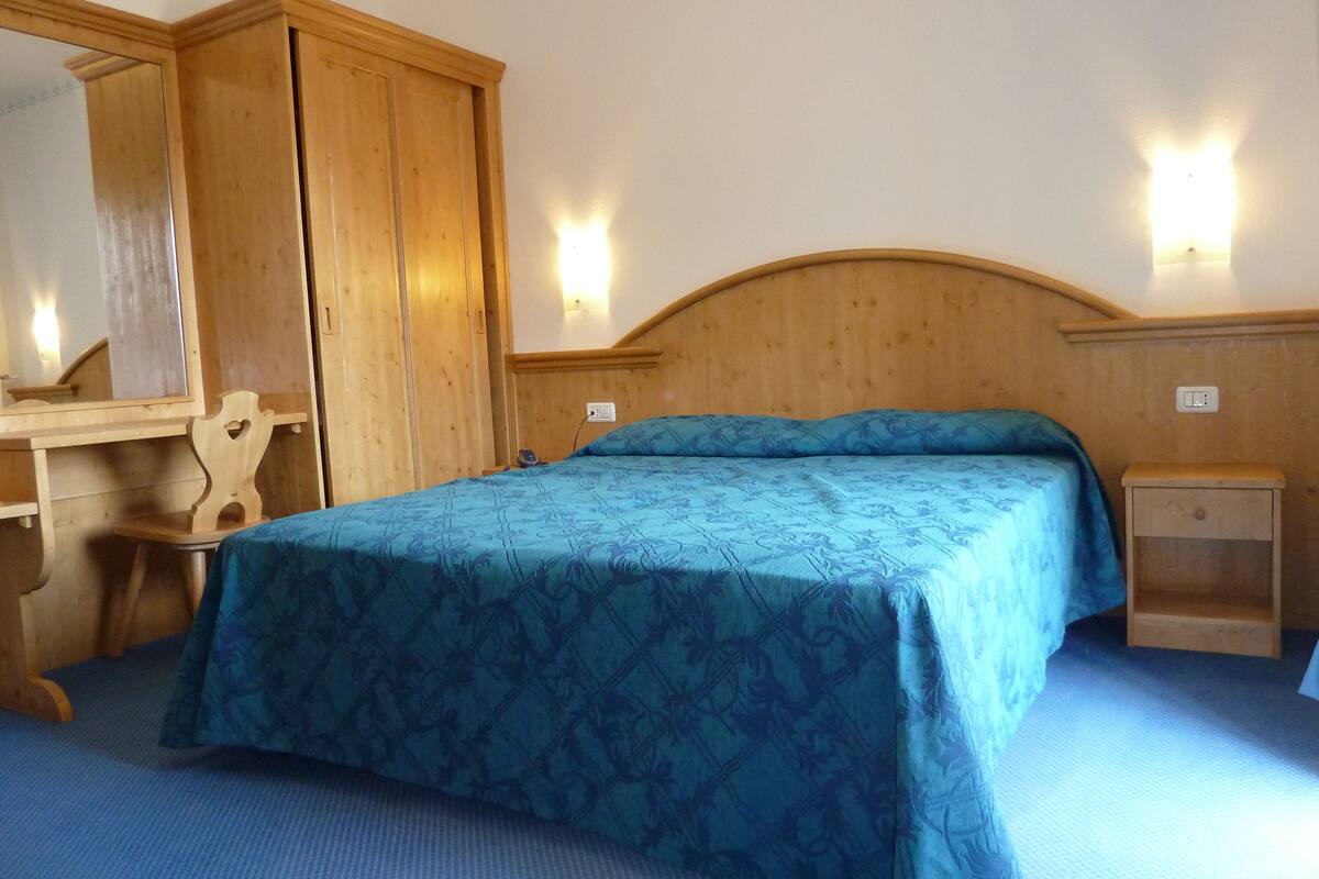 A bedroom with a wooden headboard, a bed with a blue patterned bedspread, a wooden nightstand, a mirror, and a wardrobe.
