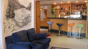 A hotel lobby with a blue sofa, three bar stools, a bar area with shelves of bottles, and a mural of a mountain landscape. - Hotel Florida (Molveno)