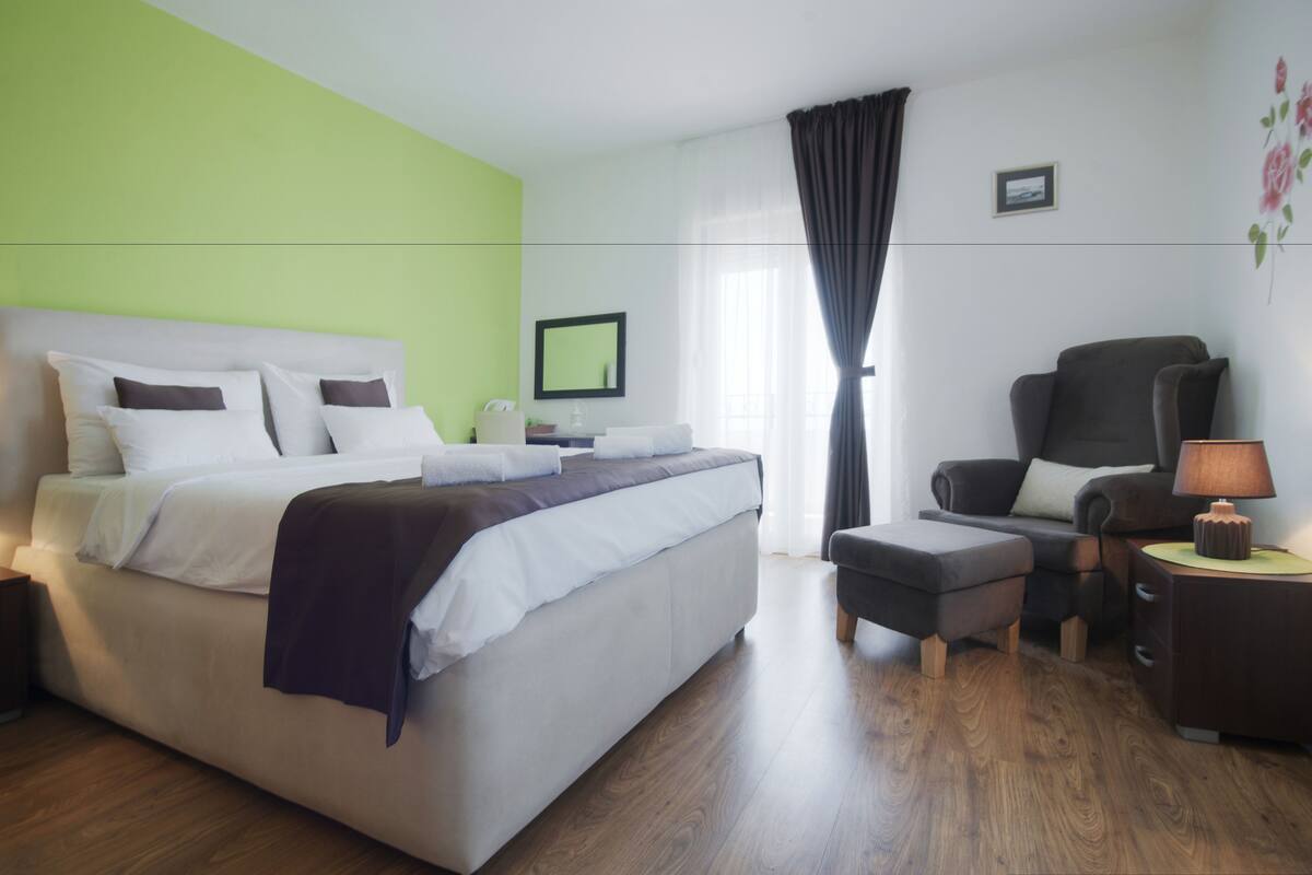 A modern hotel room with a large bed, a chair, a small table with a lamp, and a window with curtains.