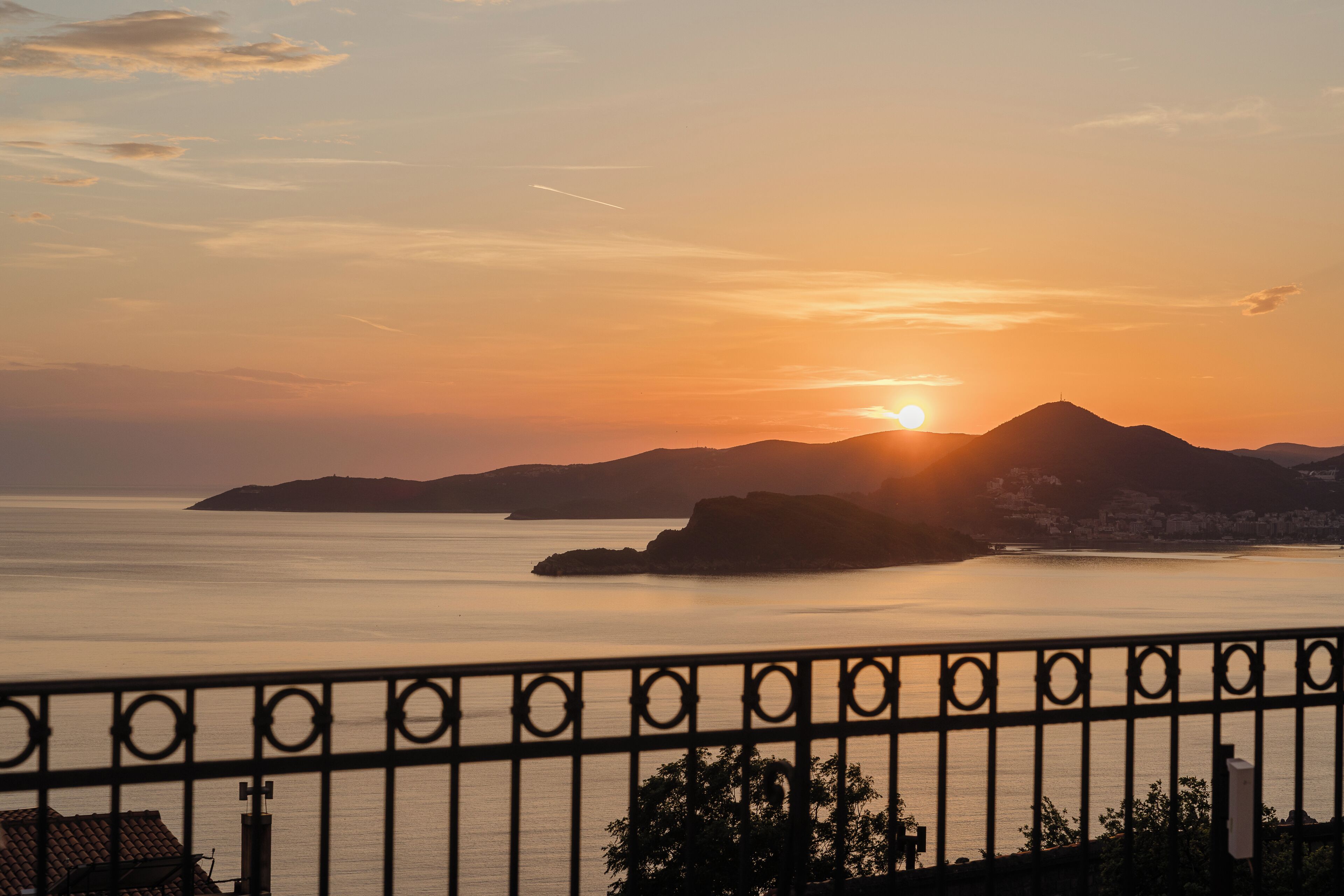 Una vista del atardecer sobre el agua, con montañas, una valla y el silueta de un edificio.