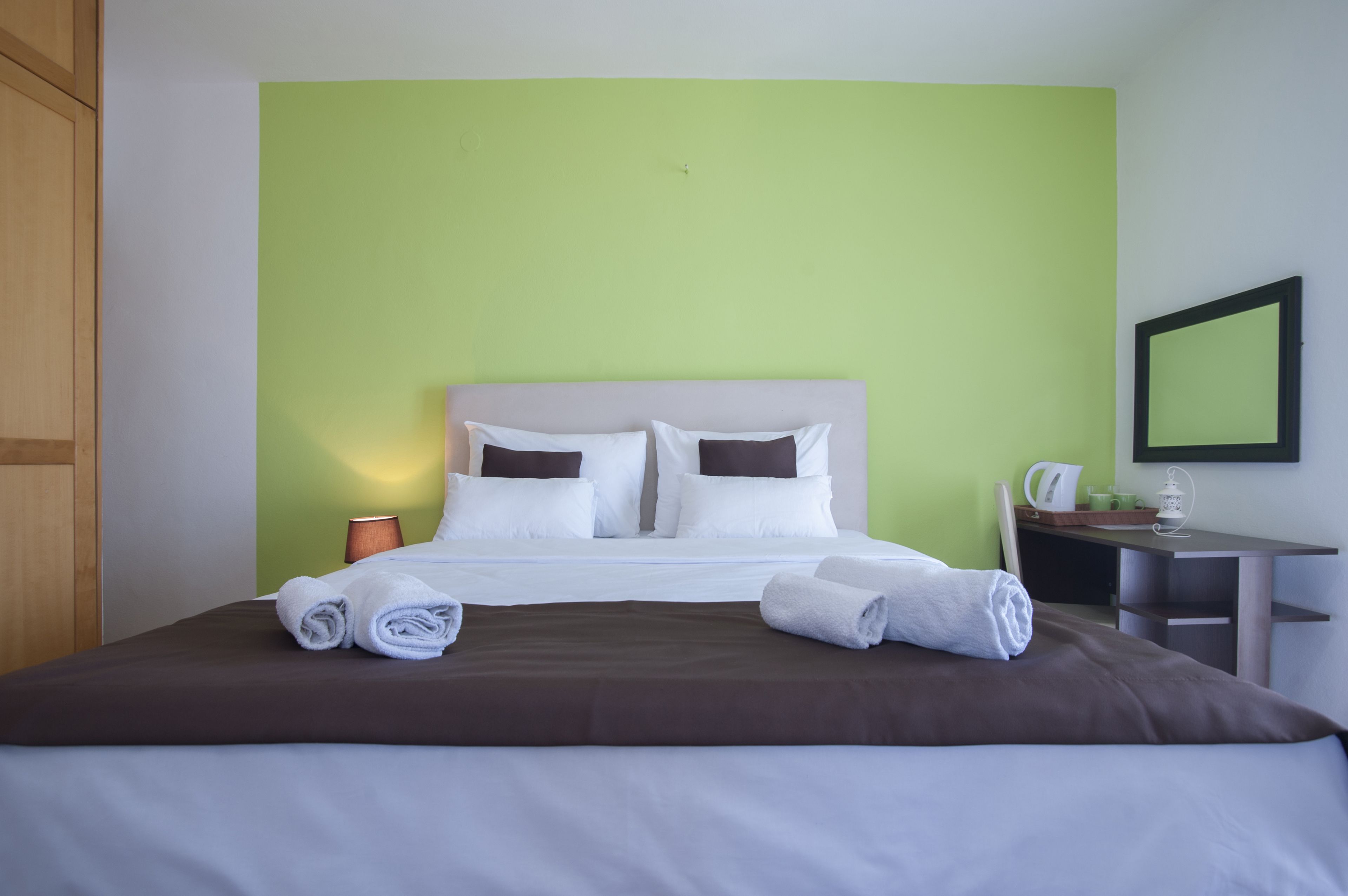 A neatly made bed with white and brown bedding, a green wall, and a small table with a lamp and a cup.