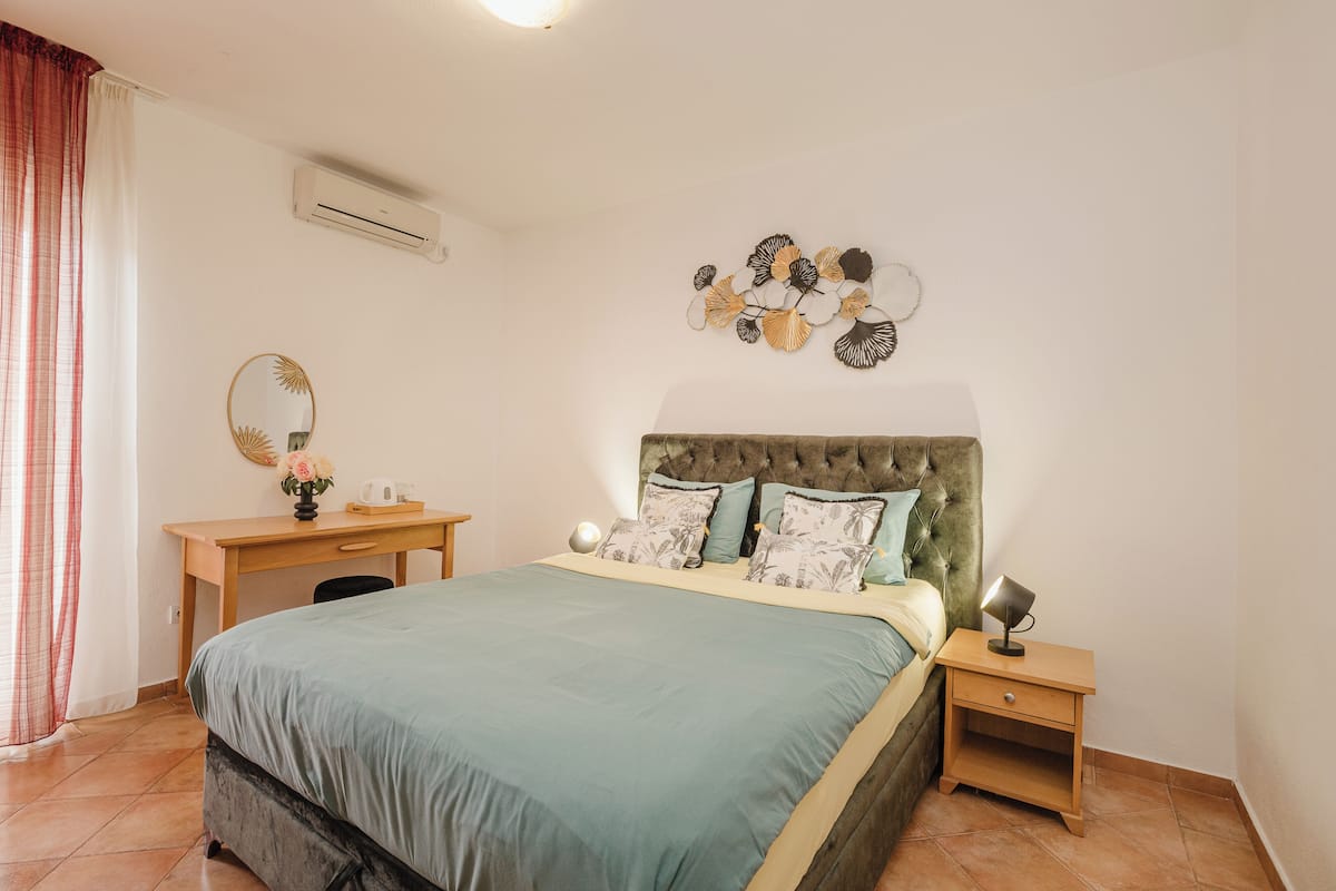 A bedroom with a bed, a desk, a chair, a nightstand, a wall-mounted air conditioner, and a decorative wall piece.