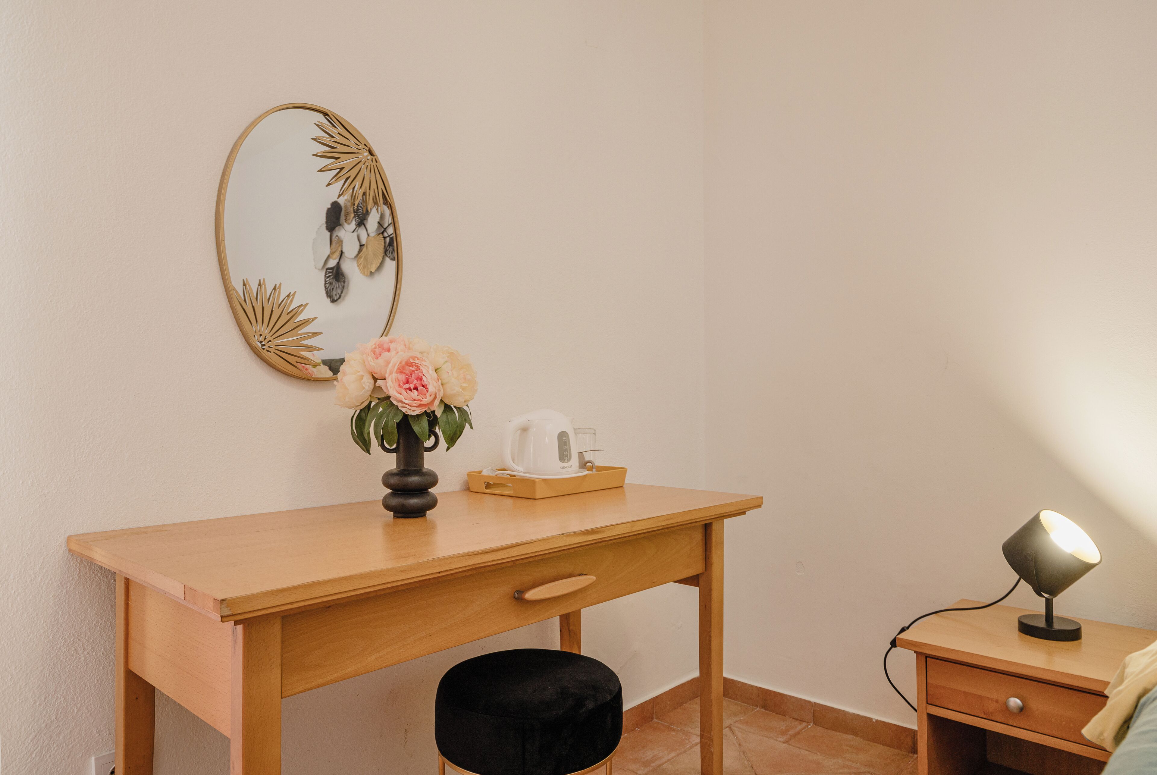 A wooden desk with a vase of flowers, a mirror, a kettle, and a lamp on a bedside table.
