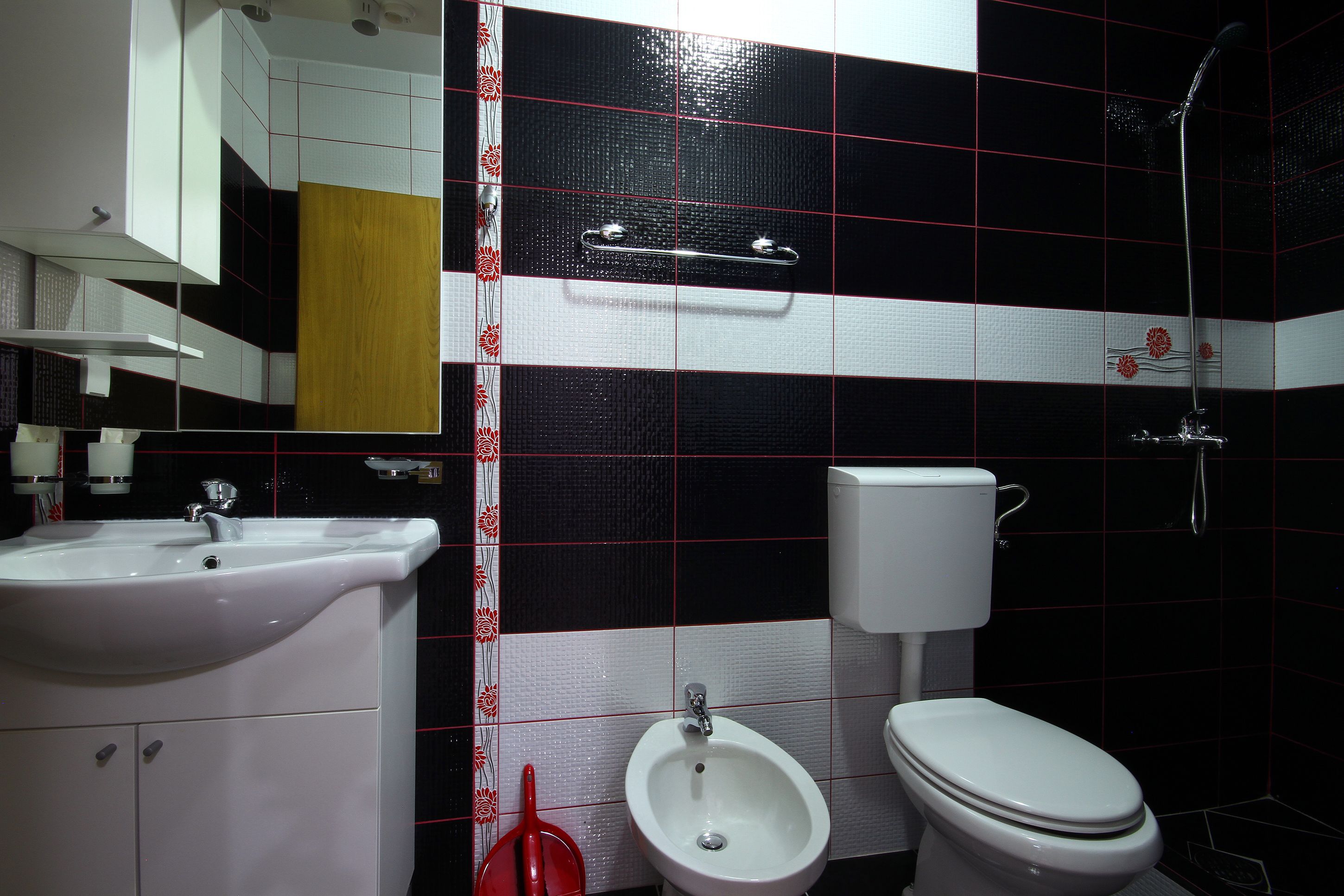 A bathroom with a white sink, bidet, and toilet, with black and white tiled walls and a red floral border.