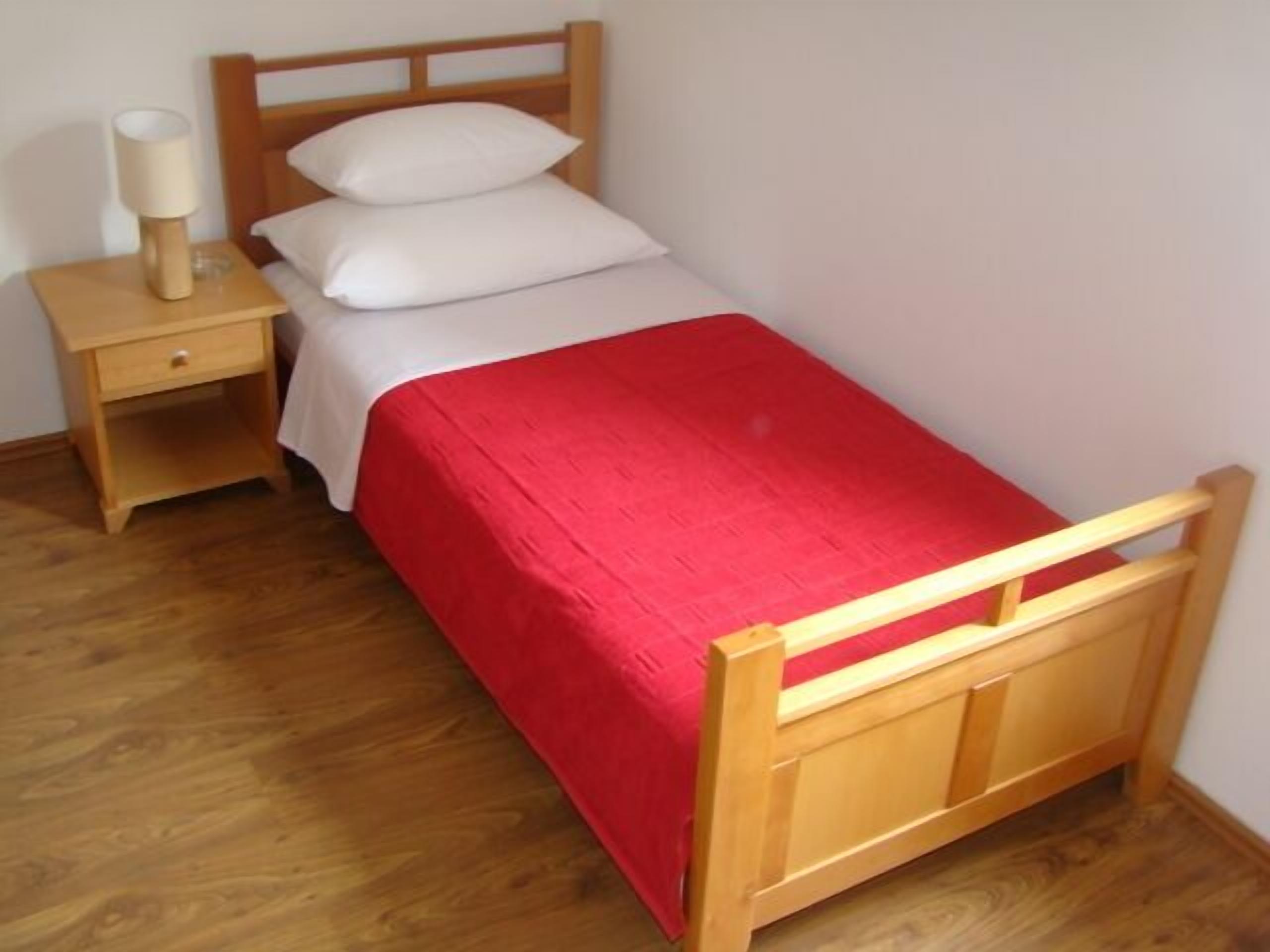 Un letto singolo con struttura in legno, copriletto rosso, due cuscini bianchi e un comodino con una lampada.