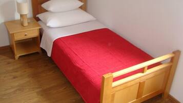 Un letto singolo con struttura in legno, copriletto rosso, due cuscini bianchi e un comodino con una lampada.