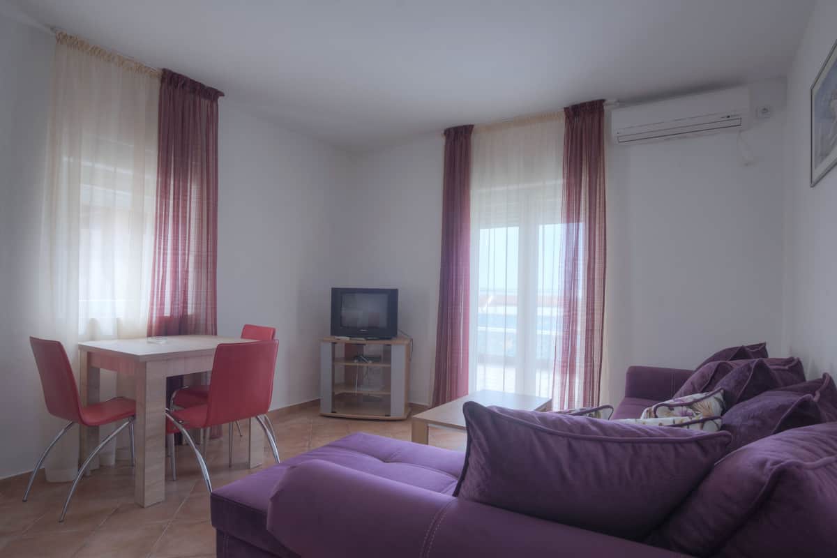 A living room with a purple sofa, a dining table with red chairs, a TV on a stand, and large windows with sheer curtains.