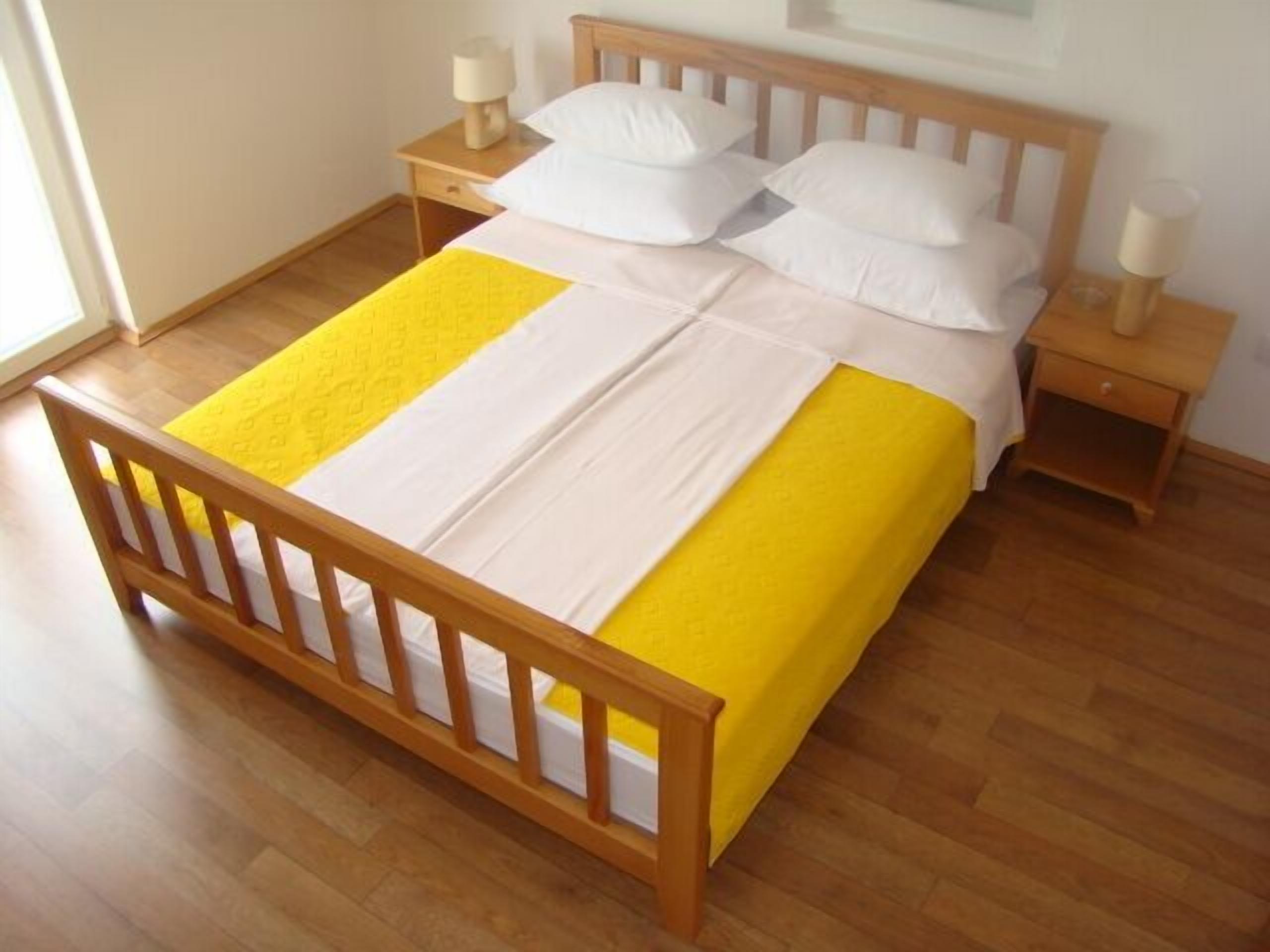 Quarto com estrutura de cama de madeira, colcha amarela e branca, almofadas brancas, duas mesas de cabeceira com abajures e piso de madeira.