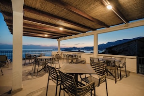 An outdoor terrace with tables and chairs, a thatched roof, and a view of the sea and mountains. - Apartments Djurasevic (Blizikuce)