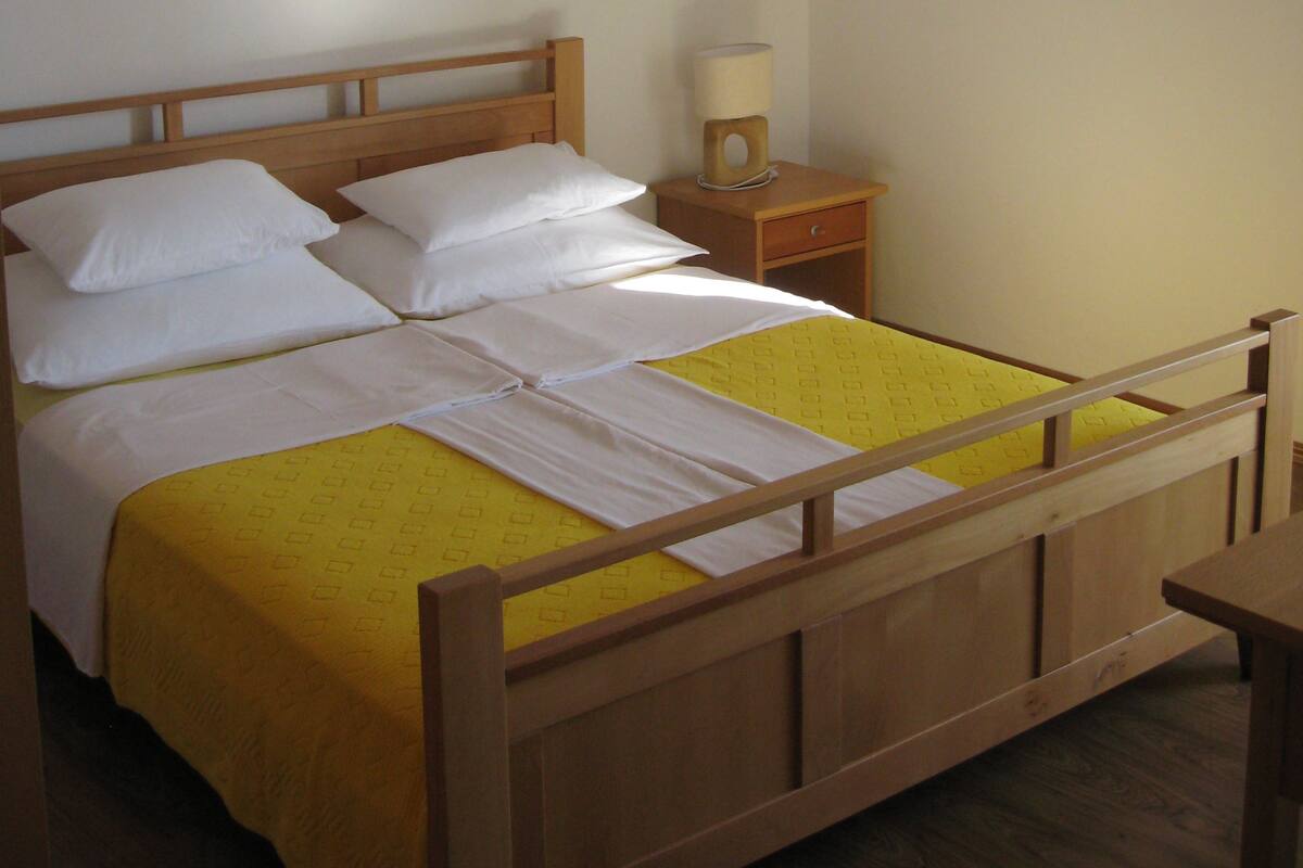 A single bed with a wooden frame, white bedding, and a yellow bedspread. There is a nightstand with a lamp and a small drawer.