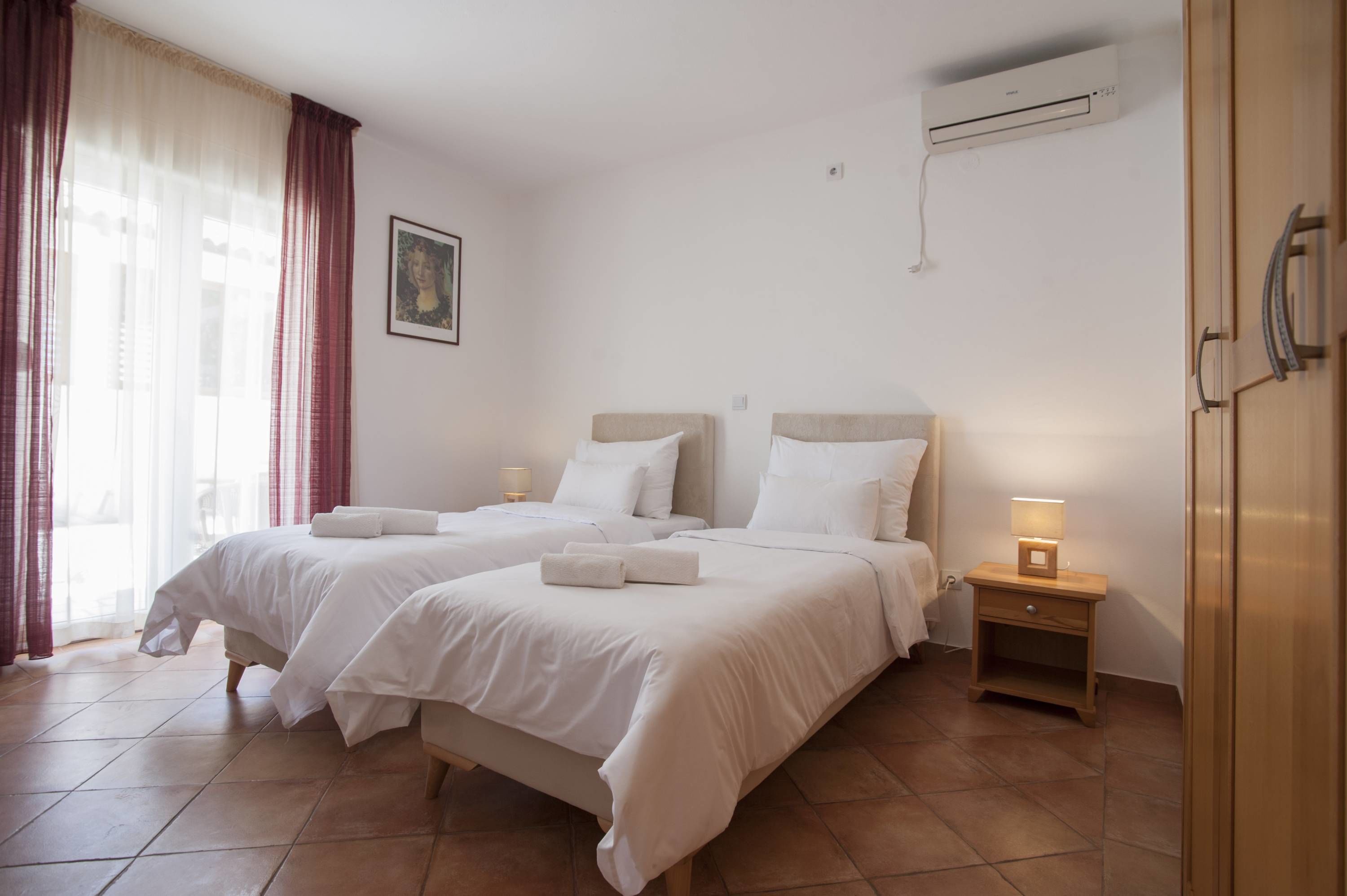 A hotel room with two beds, a nightstand with a lamp, an air conditioning unit, and a door with a handle.