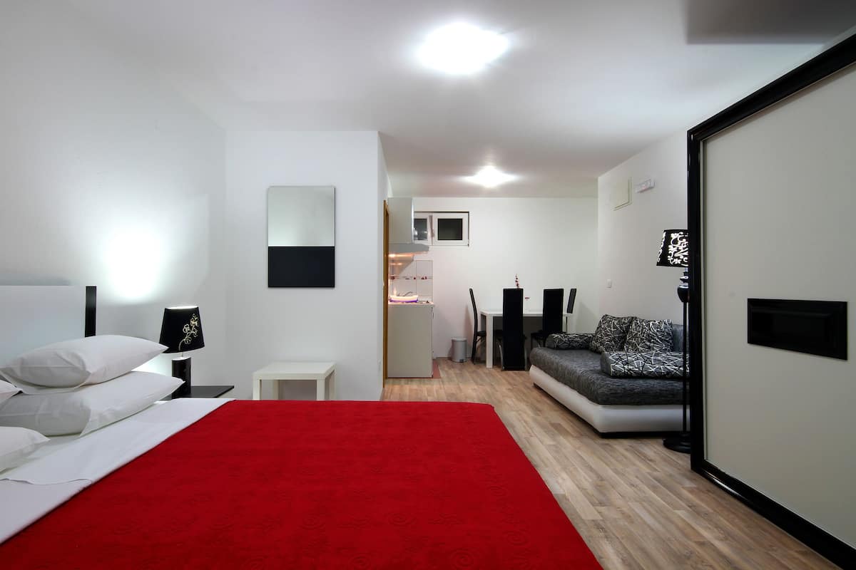 A modern hotel room with a bed, a red throw, a sofa, and a small kitchenette.