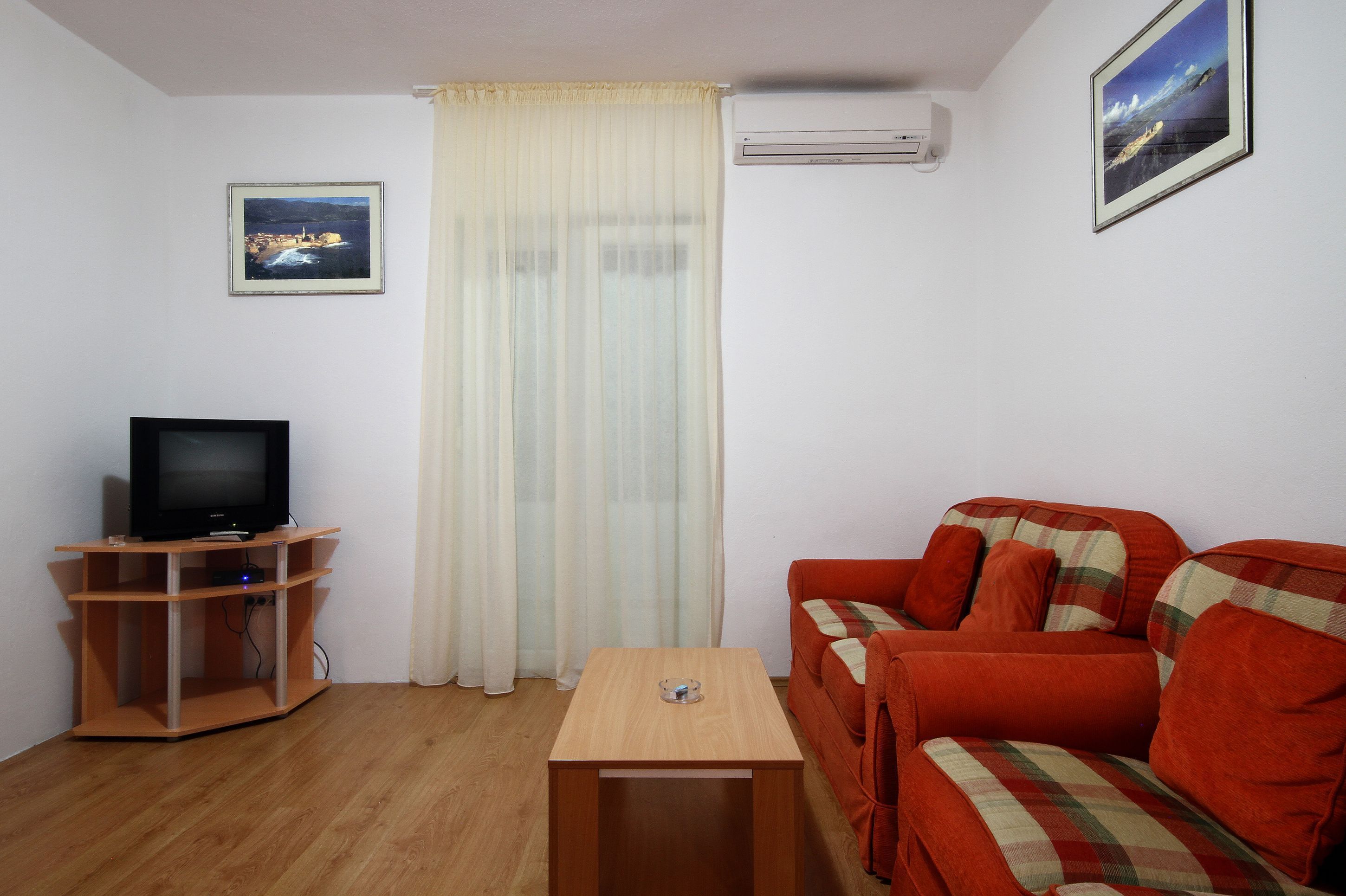 Una sala de estar con un televisor sobre un mueble de madera, un sofá rojo, una mesita de madera y una ventana con cortinas translúcidas.
