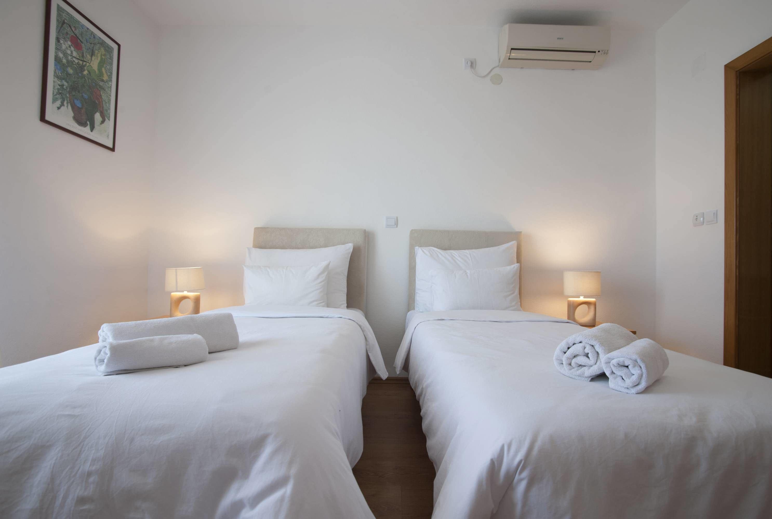 A hotel room with two beds, each with a headboard, white linens, and a rolled towel on the bed. There are bedside lamps and an air conditioning unit on the wall.