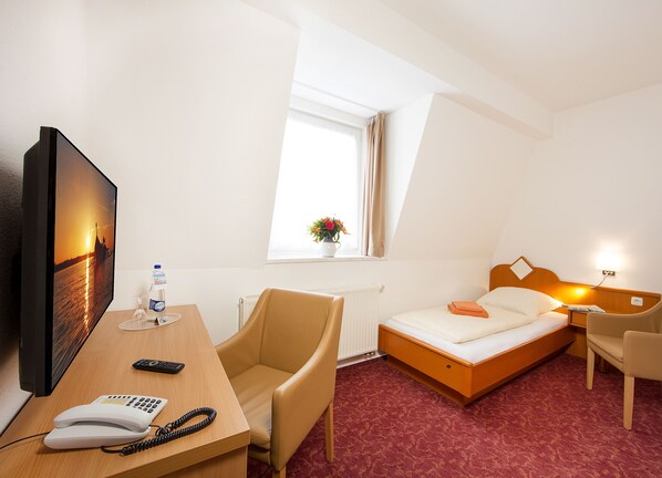 A hotel room with a bed, a desk with a telephone and remote control, a chair, a window with curtains, and a wall-mounted TV.