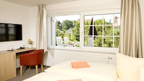 A hotel room with a bed, a desk, a chair, a TV, and a window with a view of greenery and buildings. - Ferienhotel Bernstein (Trassenheide)