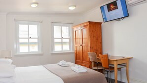 Quarto de hotel com uma cama, duas janelas, guarda-roupa de madeira, escrivaninha com cadeira e televisão na parede.
