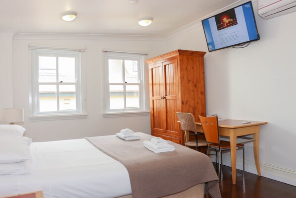 A hotel room with a bed, two windows, a wooden wardrobe, a desk with a chair, and a mounted TV.