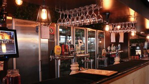Uma área de bar com várias torneiras, copos pendurados acima e uma tela de televisão exibindo uma vista urbana.