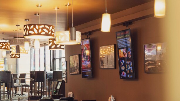 Interior de restaurante com luminárias pendentes, mesas, cadeiras e cartazes emoldurados na parede.