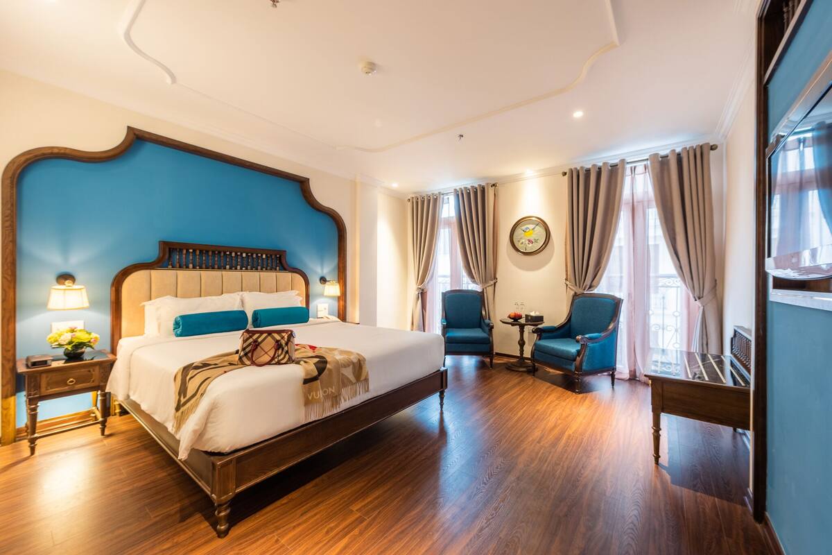 Habitación de hotel con una cama grande, cabecero de madera, una mesita pequeña, dos sillas y un ventanal con cortinas.