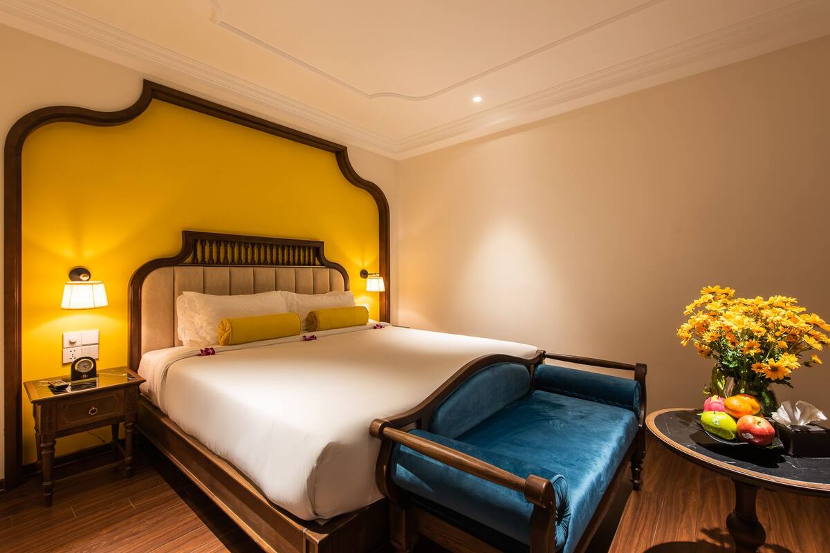 Habitación de hotel con una cama grande, una mesita de noche, un sillón azul y una pequeña mesa con un jarrón de flores.