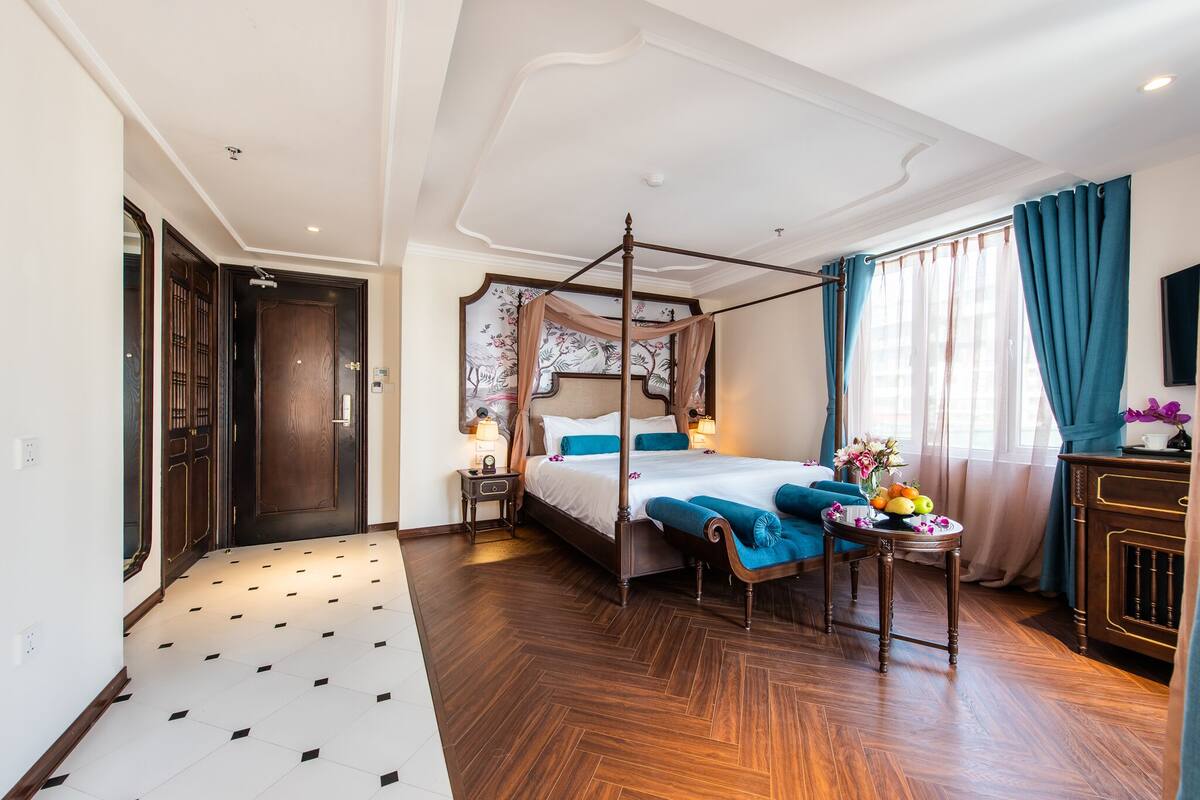 Una cama con dosel y cortinas, una mesita de noche con lámpara, y una pequeña mesa con fruta y flores.
