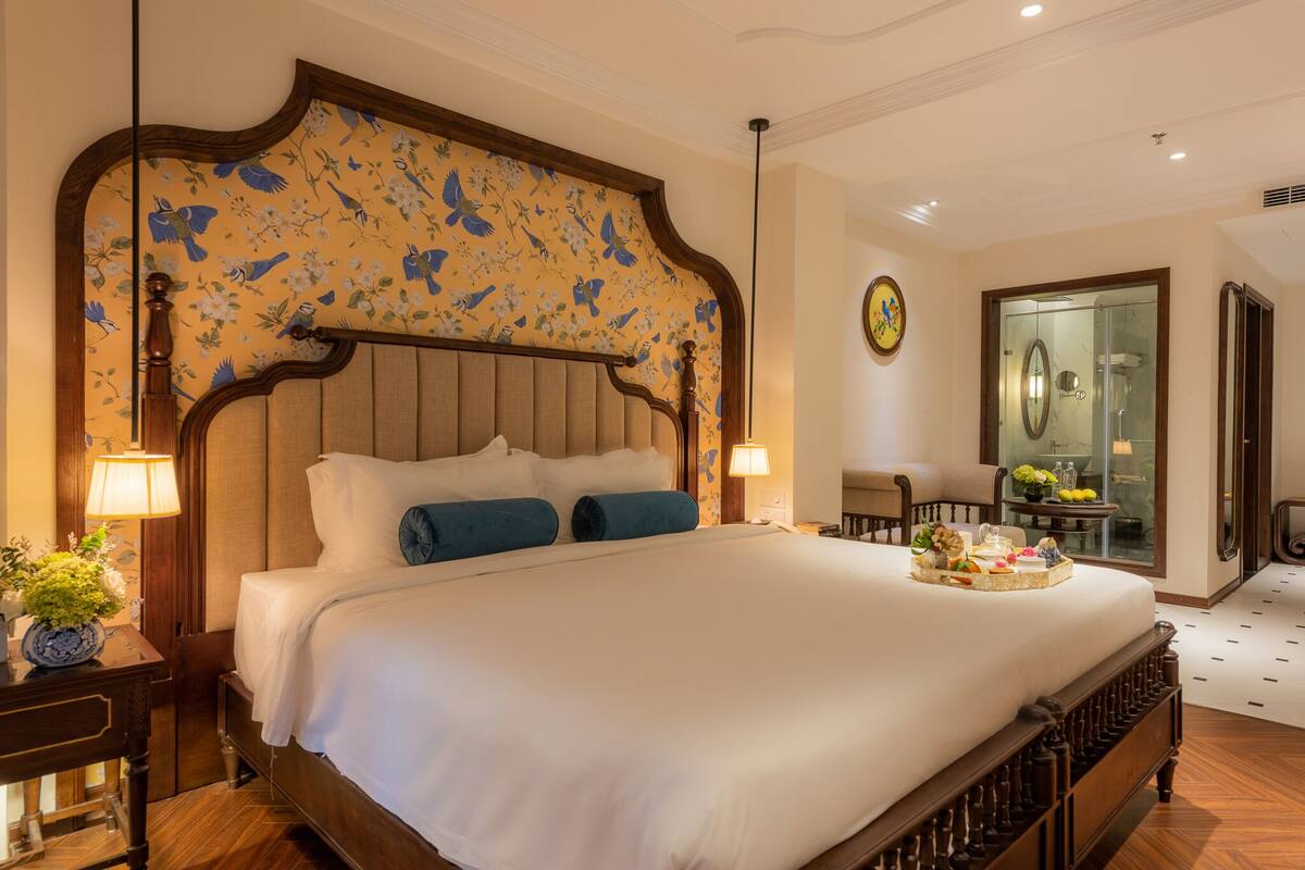 Una habitación de hotel elegante con una cama grande, cabecera de madera con papel tapiz floral, mesita de noche con lámpara y vista al baño a través de una puerta de vidrio.