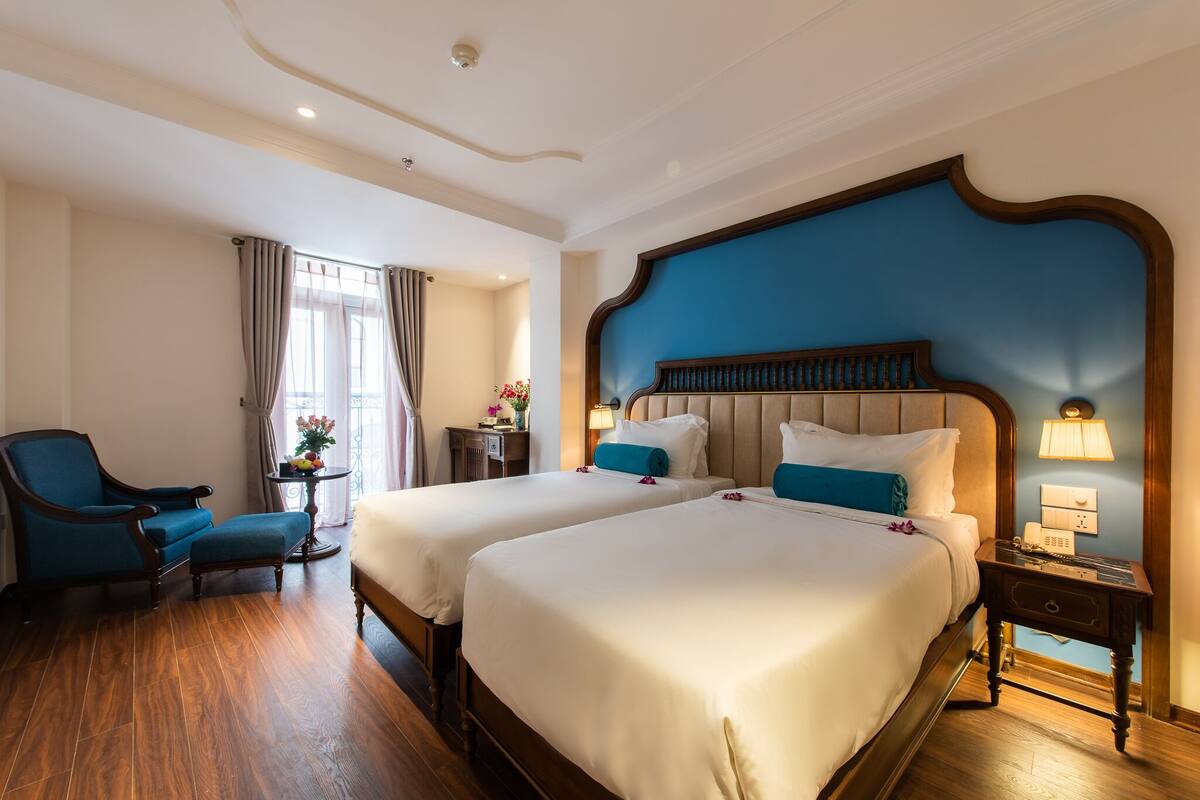Habitación de hotel con dos camas, una silla, una mesita con flores y una mesita de noche con lámpara.