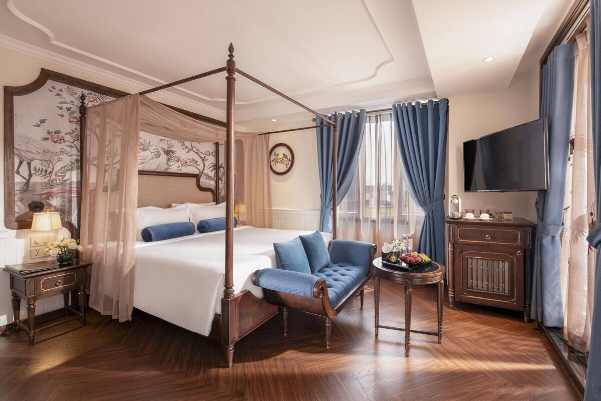Una cama con dosel y cuatro columnas, una silla, una mesita con frutero, un televisor y un reloj en la pared.